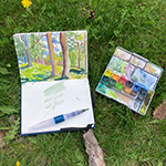 watercolour of trees and grass in a sketching type book with a paintbrush and watercolour paint palette on the grass